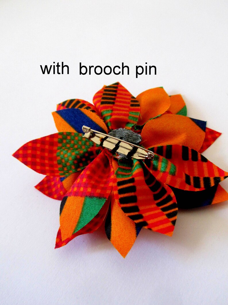 May include: A colorful fabric flower brooch with a safety pin on the back. The flower is made of orange, red, green, black, and blue fabric.