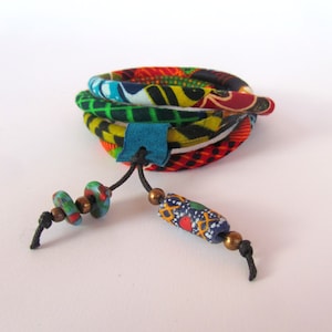 Op de afbeelding: Een kleurrijk stoffen armband met een blauwe lederen accent en een kralenkwast. De kralen zijn groen, blauw en rood met een gouden accent.