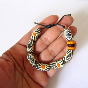Puede incluir: Un brazalete de cuentas negras y blancas con detalles naranjas. Las cuentas son cilíndricas y tienen un patrón geométrico. El brazalete está atado con un cordón negro.