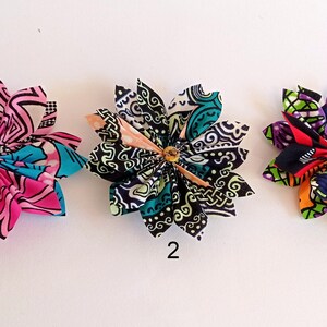 May include: Three fabric flower hair clips in different patterns. The first clip is pink, blue, and white. The second clip is black, green, and white. The third clip is red, green, purple, and orange.