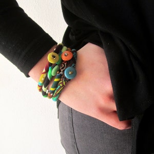 May include: A stack of colorful fabric bracelets with button accents. The bracelets are made of different patterns and colors, including green, blue, yellow, orange, and brown.
