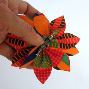 May include: A colorful fabric flower brooch with a pin backing. The flower has petals in shades of orange, red, green, blue, and black.