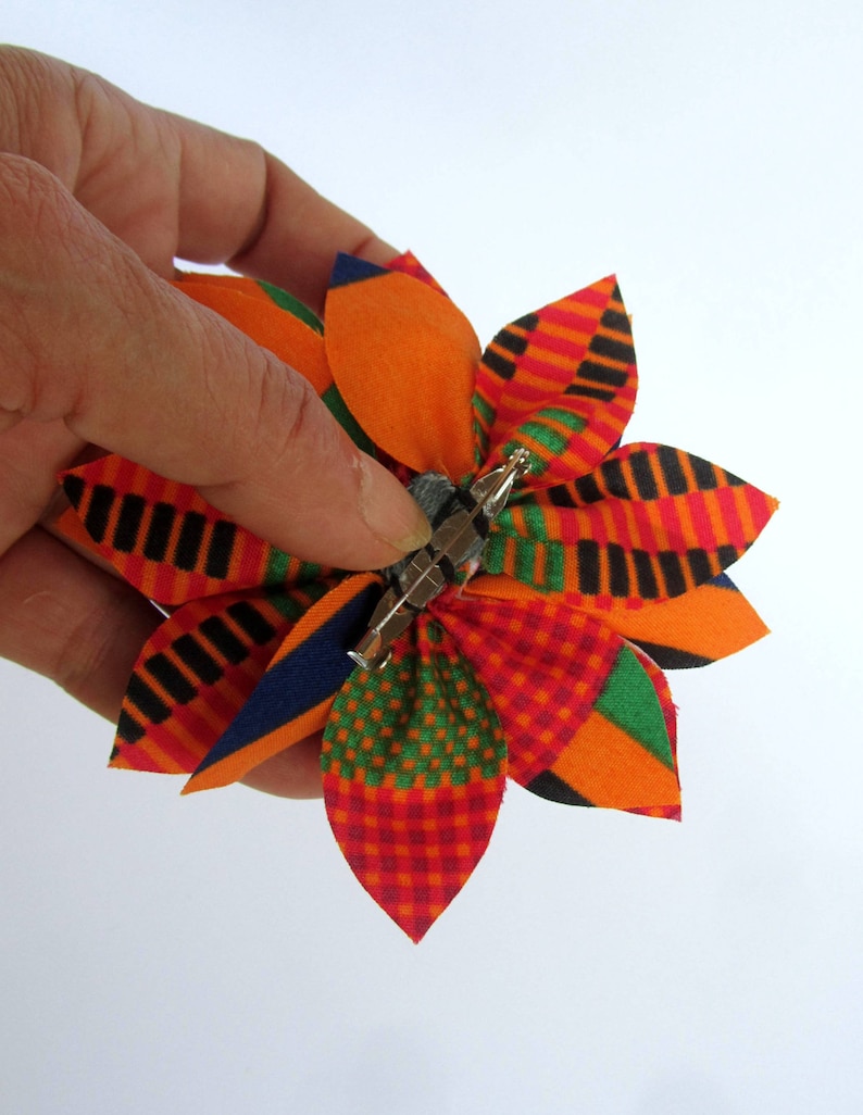 May include: A colorful fabric flower brooch with a pin backing. The flower has petals in shades of orange, red, green, blue, and black.