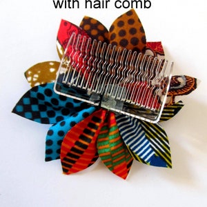 May include: A colorful fabric flower hair clip with a clear plastic comb attached. The flower is made of multiple fabric petals in various patterns, including polka dots, stripes, and geometric designs. The colors include red, orange, yellow, green, blue, and black.