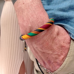 Puede incluir: Un brazalete tejido colorido con rayas rojas, amarillas, verdes y negras. El brazalete está atado con un cordón negro y tiene una pequeña cuenta dorada.