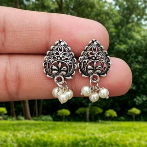 May include: A pair of silver-toned earrings with a filigree design and small dangling faux pearls. The earrings have a teardrop shape and are held up by a person's fingers against a blurred green background.