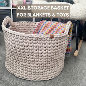 May include: A large, beige, woven storage basket with handles, holding a colorful blanket. The text "XXL STORAGE BASKET FOR BLANKETS & TOYS" is displayed across the top. A small, brown tag is attached to the basket.