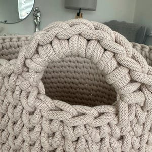 May include: Close-up of a beige, hand-knitted basket with a large, braided handle. The basket's body features a textured, open weave, and the handle is thick and sturdy. The neutral color and chunky knit suggest a cozy, handmade aesthetic, suitable for home decor.