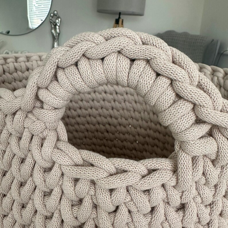 May include: Close-up of a beige, hand-knitted basket with a large, braided handle. The basket's body features a textured, open weave, and the handle is thick and sturdy. The neutral color and chunky knit suggest a cozy, handmade aesthetic, suitable for home decor.