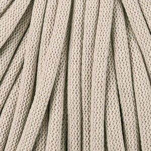 May include: Close-up of beige macrame cord, tightly woven with a textured, ridged pattern. The cord is arranged in a series of parallel lines, creating a sense of depth and texture. Ideal for crafting and home decor projects.