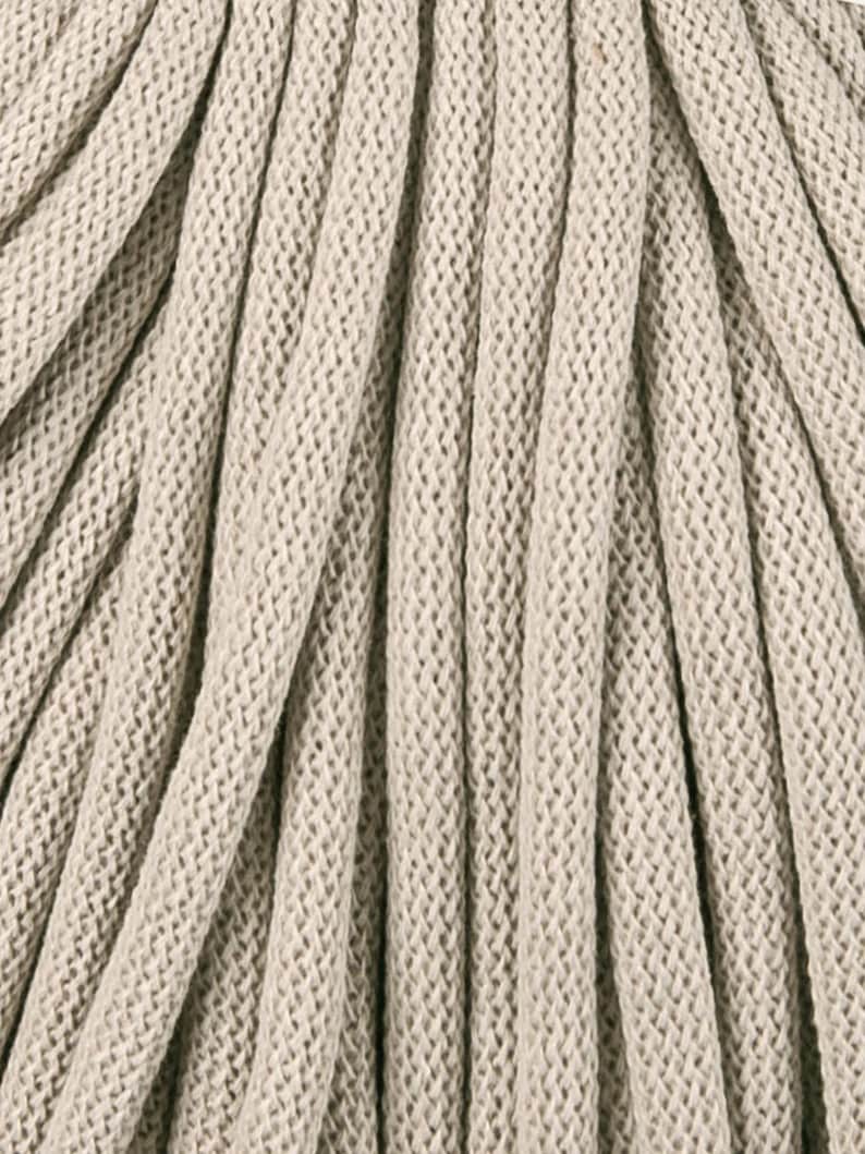May include: Close-up of beige macrame cord, tightly woven with a textured, ridged pattern. The cord is arranged in a series of parallel lines, creating a sense of depth and texture. Ideal for crafting and home decor projects.