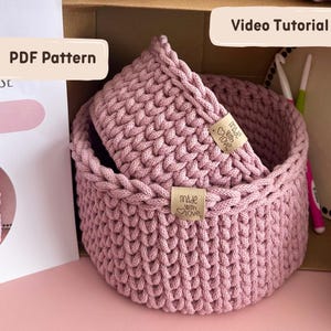 May include: Two handmade, dusky pink crochet baskets, one nested inside the other. Each basket has a woven texture and a "made with love" tag. The image also includes text that says "PDF Pattern" and "Video Tutorial".