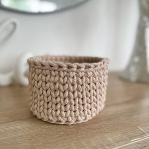Può includere: Un piccolo cestino rotondo all'uncinetto beige. Il cestino è realizzato con filato spesso, con un motivo a punto strutturato. Il bordo del cestino ha un bordo intrecciato. Il cestino è appoggiato su una superficie di legno.