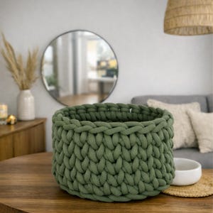 K&ouml;nnte beinhalten: Ein olivgr&uuml;ner Strickkorb aus dickem Garn steht auf einem Holztisch. Der Korb hat ein strukturiertes Aussehen und ist leer. Der Korb hat einen Durchmesser von ca. 25 cm. Wohnkultur.