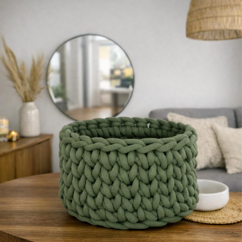 K&ouml;nnte beinhalten: Ein olivgr&uuml;ner Strickkorb aus dickem Garn steht auf einem Holztisch. Der Korb hat ein strukturiertes Aussehen und ist leer. Der Korb hat einen Durchmesser von ca. 25 cm. Wohnkultur.