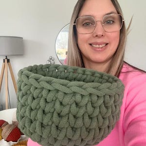 K&ouml;nnte beinhalten: Ein handgeh&auml;kelter Korb in Salbeigr&uuml;n. Der Korb ist rund mit einer geflochtenen Textur und einer breiten &Ouml;ffnung. Das Garn scheint ein dickes, weiches Material zu sein. Der Korb wird vor einer Person gehalten, die ein rosa Oberteil tr&auml;gt.