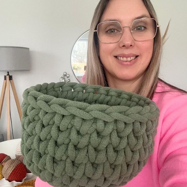K&ouml;nnte beinhalten: Ein handgeh&auml;kelter Korb in Salbeigr&uuml;n. Der Korb ist rund mit einer geflochtenen Textur und einer breiten &Ouml;ffnung. Das Garn scheint ein dickes, weiches Material zu sein. Der Korb wird vor einer Person gehalten, die ein rosa Oberteil tr&auml;gt.