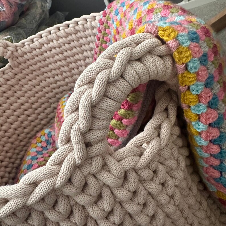 May include: Close-up of a beige knitted basket with braided handles. The basket is filled with a colorful, crocheted blanket in shades of pink, blue, yellow, and green. The basket's texture is visible.