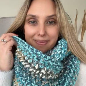 May include: A chunky knit infinity scarf in shades of teal, gray, white, and tan. The scarf is made of thick yarn and has a textured appearance. The scarf is wrapped around the neck.