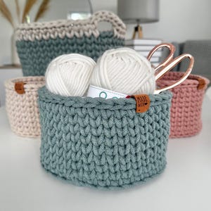 以下が含まれることがあります： さまざまなサイズと色の手作りのかぎ針編みバスケットのコレクション。手前のバスケットはティール色で、白い糸と「MADE WITH」と書かれたタグが入っています。他のバスケットは、ライトピンク、ベージュ、ティール色で、ハンドルが付いています。