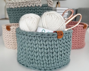 May include: A collection of handmade crochet baskets in various sizes and colors. The foreground basket is teal and filled with white yarn and a tag that says "MADE WITH". Other baskets are light pink, beige, and teal with a handle.