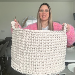 May include: A large, off-white, hand-knitted basket with two sturdy handles. The basket is made of thick, chunky yarn, creating a textured surface. The basket is designed for storage and organization.