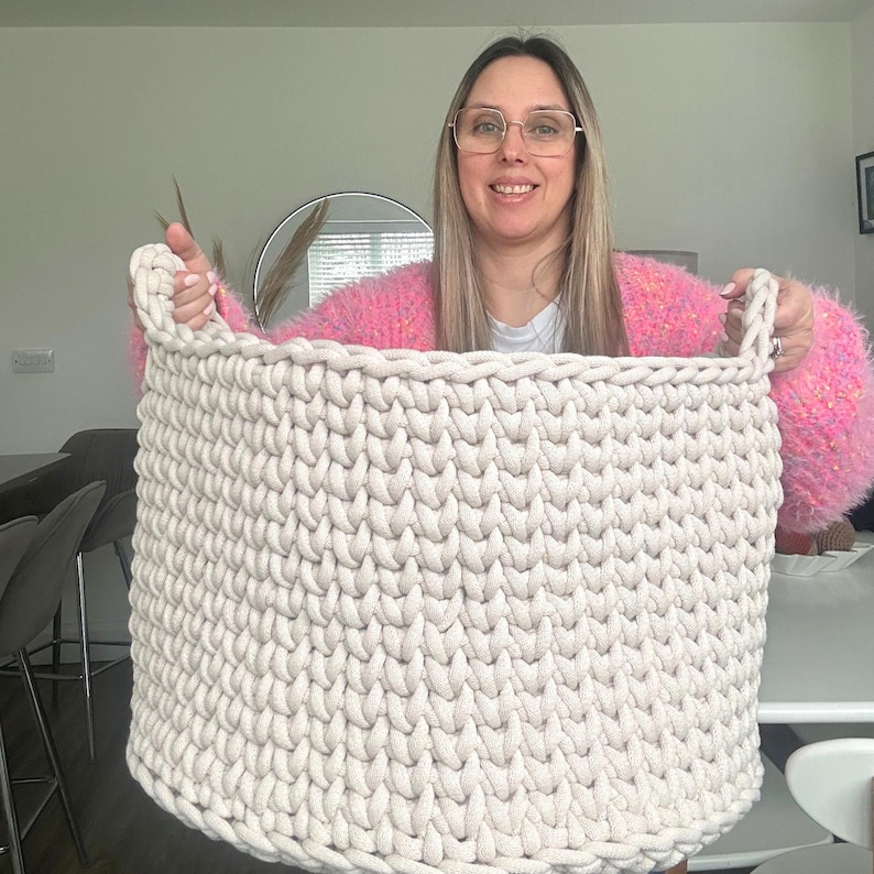 May include: A large, off-white, hand-knitted basket with two sturdy handles. The basket is made of thick, chunky yarn, creating a textured surface. The basket is designed for storage and organization.