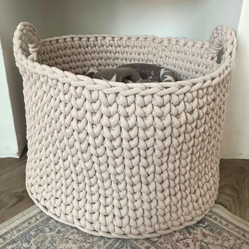 May include: A large, beige, hand-crocheted basket with two handles. The basket is filled with a folded blanket. The basket is made of thick, soft yarn and has a textured appearance. The basket is sitting on a patterned rug.