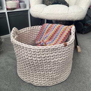 May include: A large, beige, hand-knitted storage basket with two handles. The basket contains a colorful, square, crocheted blanket. The basket is made of thick yarn and has a textured appearance. A small, brown tag is attached to the side.