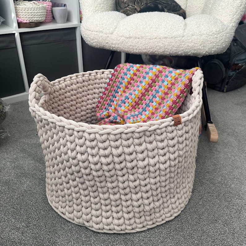 May include: A large, beige, hand-knitted storage basket with two handles. The basket contains a colorful, square, crocheted blanket. The basket is made of thick yarn and has a textured appearance. A small, brown tag is attached to the side.