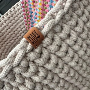 May include: Close-up of a beige, hand-knitted basket with a braided design. A brown leather tag with the words "MADE WITH" and a heart is attached to the basket. A colorful, crocheted blanket is visible inside the basket.