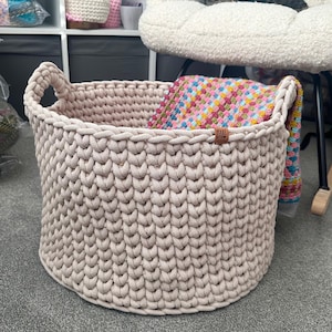 May include: A large, beige, hand-knitted basket with two handles. The basket is filled with a colorful, patterned blanket. A small, brown tag with the word "MADE" is attached to the basket.