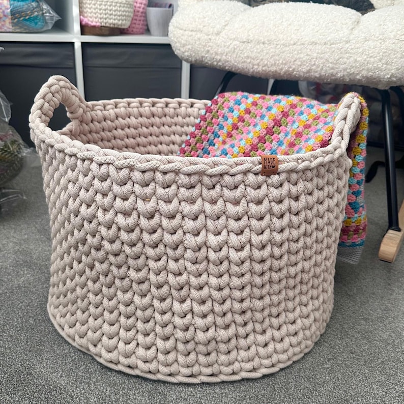 May include: A large, beige, hand-knitted basket with two handles. The basket is filled with a colorful, patterned blanket. A small, brown tag with the word "MADE" is attached to the basket.