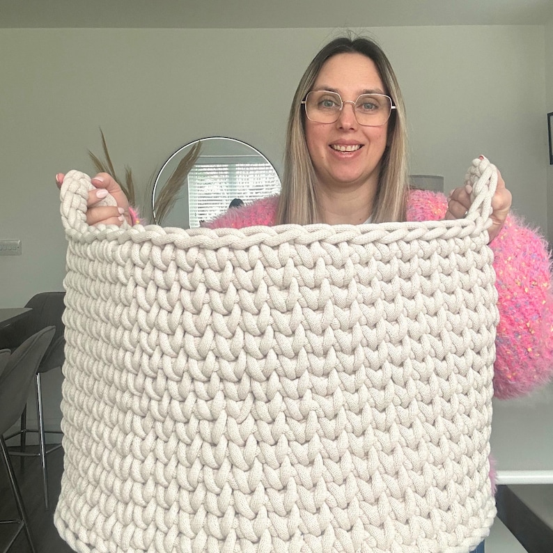 May include: A large, rectangular, off-white crochet basket with two handles. The basket is made of thick, textured yarn and appears sturdy. The neutral color makes it suitable for various home decor styles. The basket is held up by a person.