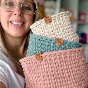 以下が含まれることがあります： クリーム色、ティール、ピンクの3つのかぎ針編みバスケットが積み重ねられています。各バスケットには「MADE WITH」と書かれた革製のタグが付いています。バスケットは太い糸でできており、質感があります。バスケットは手作りで、収納に適しています。