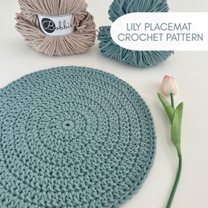 Peut inclure: Nappe crochetée de couleur vert d'eau, avec un motif circulaire. L'image présente également deux pelotes de fil, une beige et une turquoise, et une tulipe. Le texte "LILY PLACEMAT CROCHET PATTERN" est visible.