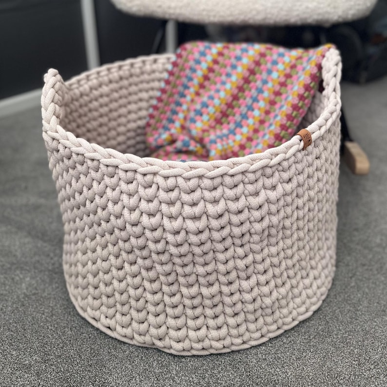 May include: A beige, round, crocheted basket with two handles. The basket is filled with a colorful, patterned blanket. A small, brown leather tag is attached to the side of the basket.