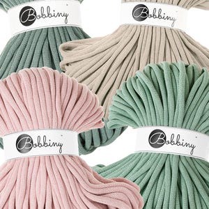 K&ouml;nnte beinhalten: Nahaufnahme von vier Kn&auml;ueln Bobbiny Makramee-Garn in Pastellfarben: Salbeigr&uuml;n, Beige, Hellrosa und Mintgr&uuml;n. Jedes Kn&auml;uel ist mit einem wei&szlig;en Etikett mit dem Bobbiny-Logo in schwarzer Schrift umwickelt.