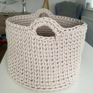 May include: A beige, hand-crocheted storage basket with two sturdy handles. The basket has a rounded shape and a textured surface created by the tight crochet stitches. The neutral color and simple design make it suitable for various home decor styles.