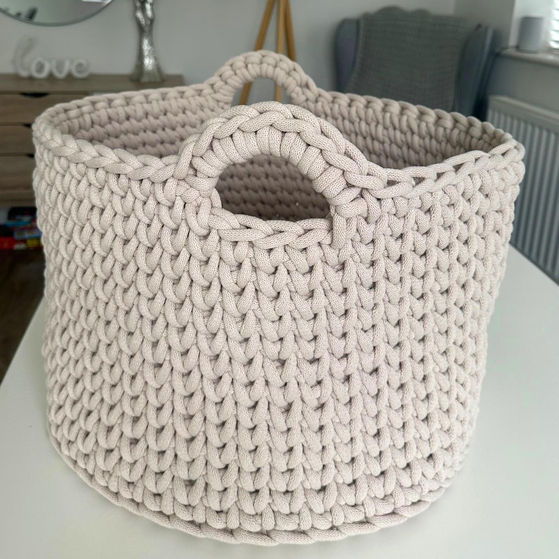 May include: A beige, hand-crocheted storage basket with two sturdy handles. The basket has a rounded shape and a textured surface created by the tight crochet stitches. The neutral color and simple design make it suitable for various home decor styles.