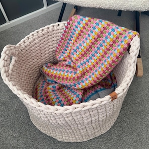May include: A large, beige, woven basket with two handles, filled with a colorful, crocheted blanket. The blanket features a patchwork design with pink, blue, yellow, and green squares. A small, brown tag is attached to the basket.