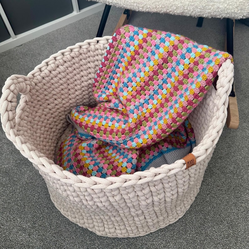 May include: A large, beige, woven basket with two handles, filled with a colorful, crocheted blanket. The blanket features a patchwork design with pink, blue, yellow, and green squares. A small, brown tag is attached to the basket.