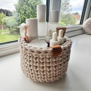 May include: A beige, hand-crocheted basket filled with skincare products. The basket has a textured, woven design and a small leather tag that reads "MADE WITH". The products include white bottles, droppers, and a rose gold facial roller.