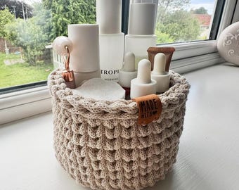 May include: A beige, hand-crocheted basket filled with skincare products. The basket has a textured, woven design and a small leather tag that reads "MADE WITH". The products include white bottles, droppers, and a rose gold facial roller.