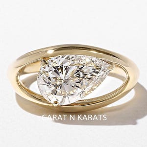 May include: A gold ring featuring a pear-shaped diamond. The diamond is set within a gold frame, creating a unique and modern design. The ring is displayed on a white surface, highlighting its brilliance. The ring is from Carat N Karats.