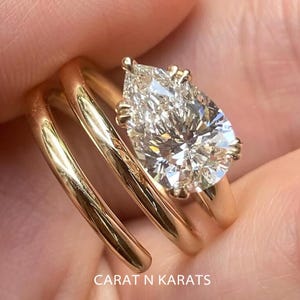 May include: A gold ring with a pear-shaped diamond. The ring has a unique spiral design. The text "CARAT N KARATS" is visible on the ring. The diamond is clear and sparkling.
