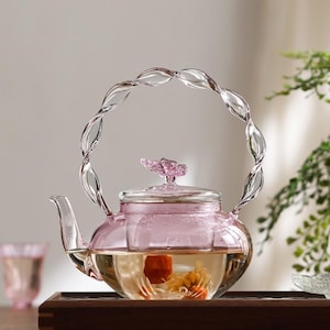 Puede incluir: Una tetera de vidrio transparente con un tinte rosado, llena de té y una flor. La tetera tiene un asa transparente y retorcida y una tapa en forma de flor. La tetera está sobre una bandeja de madera.