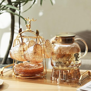 May include: An elegant tea set comprising six amber-coloured glass cups and saucers on a gold-coloured metal rack. A matching glass teapot with a gold lid and handle is also present. The set is displayed on a clear tray.