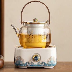 Op de afbeelding: Een heldere glazen theepot met een bruine handgreep en een witte keramische basis. De theepot bevat een gouden vloeistof en staat op een witte warmer met blauwe golfontwerpen. De deksel heeft een decoratieve knop.