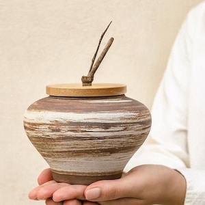 Puede incluir: Un tarro de cerámica artesanal con tapa de madera y asa de ramita. El tarro presenta bandas horizontales marrones y blancas, creando una estética texturizada y terrosa. El tarro está sostenido en las manos.